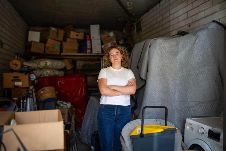 Sarah, wears a white tshirt and jeans standing among her boxed up belongings inside a garage